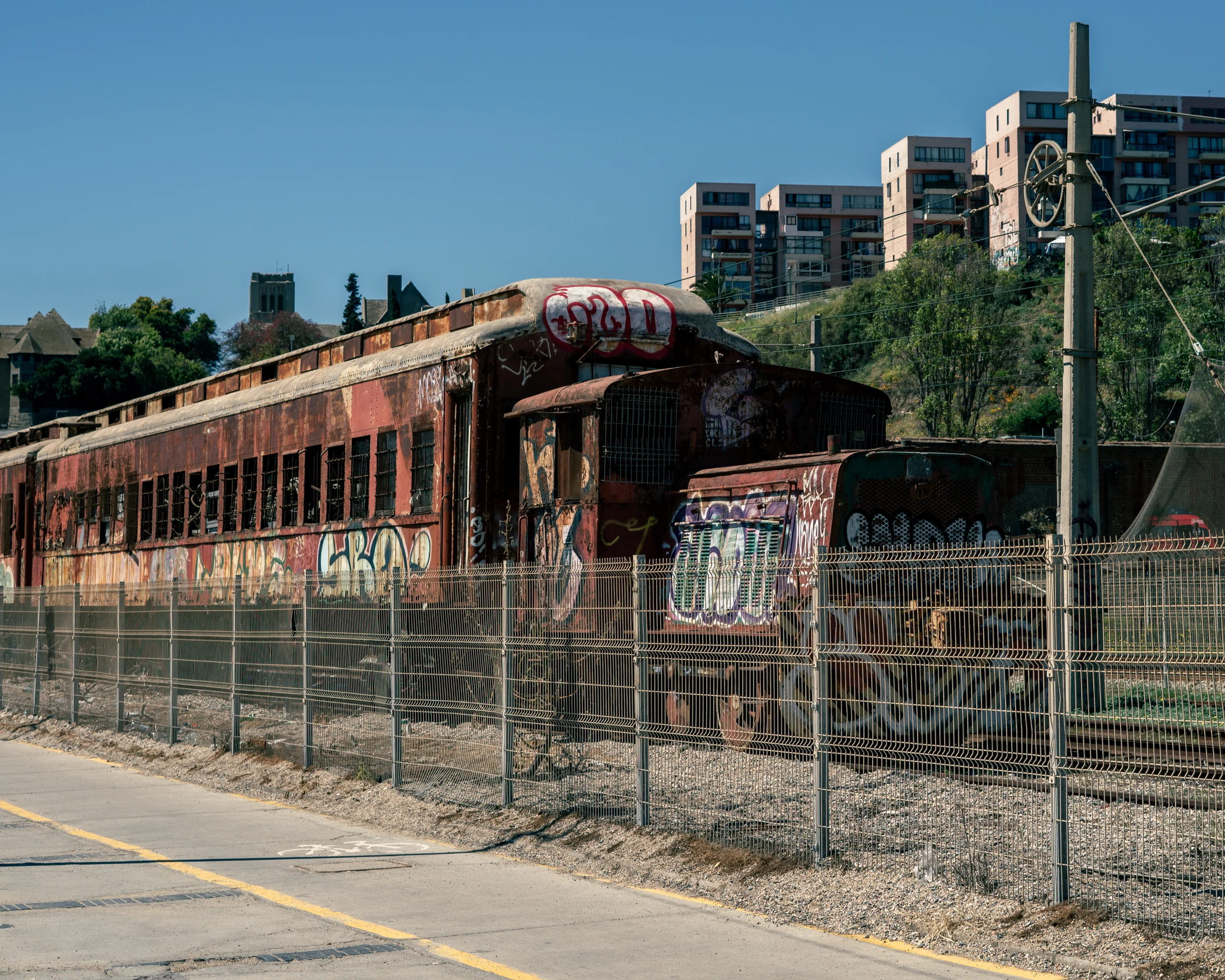 Tren abandonado detrás de una reja, cubierto de grafitis, detenido hace rato pero todavía presente en el paisaje.