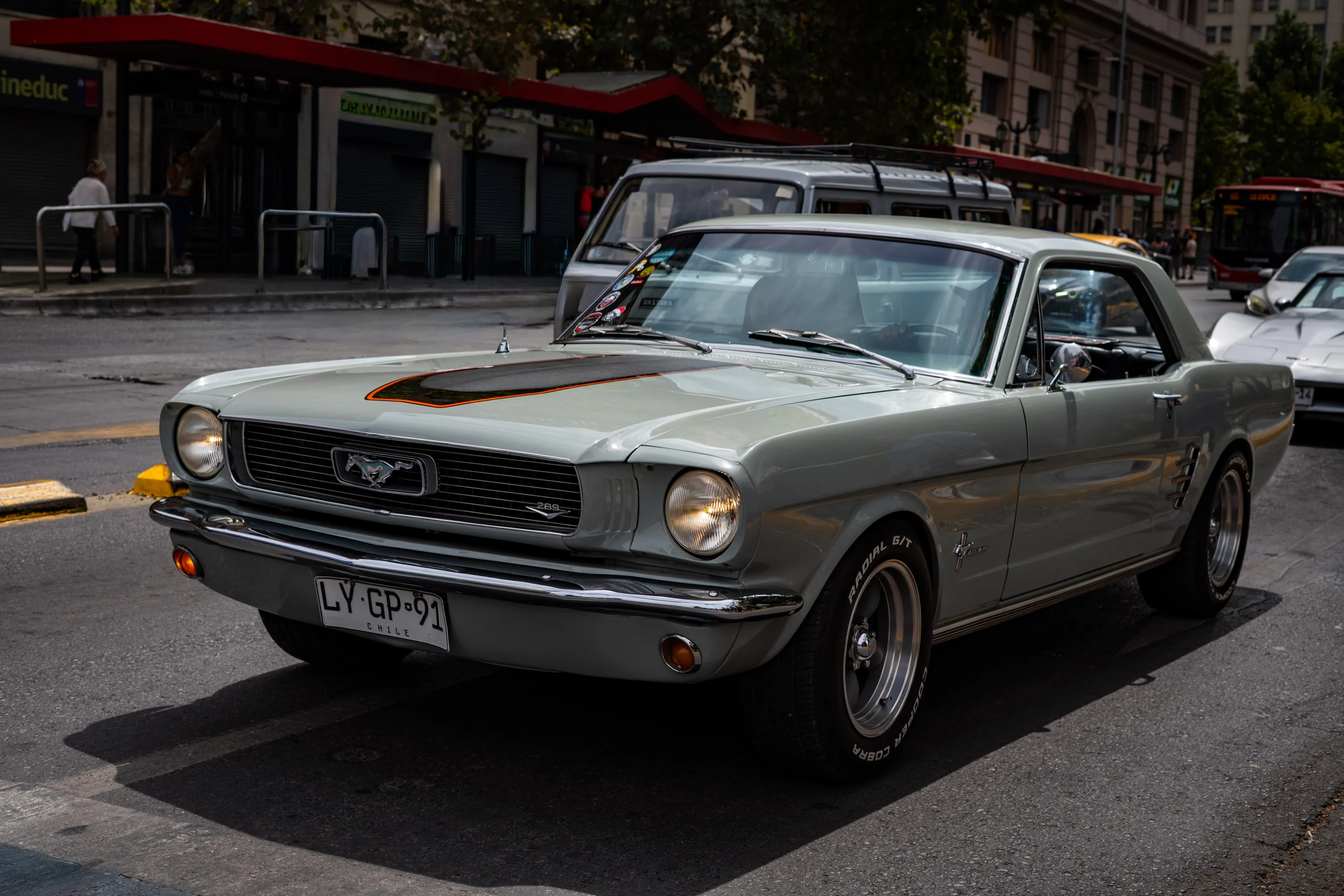 Automóvil Ford Mustang clásico color gris con detalles en negro y naranja estacionado en una calle urbana. Se observan edificios y otros vehículos al fondo, junto con algunos peatones. El automóvil tiene placas de Chile y neumáticos radiales.