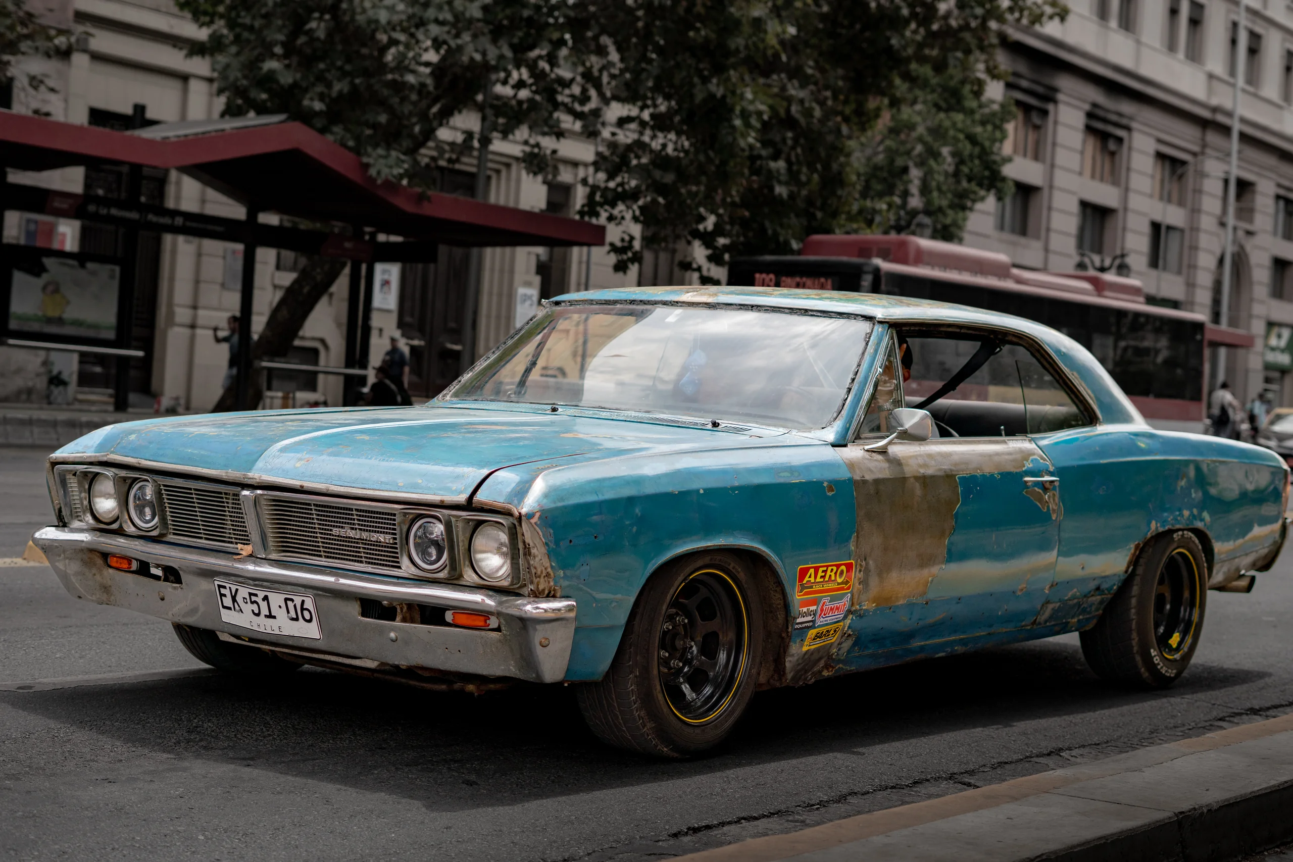 Imagen de un automóvil clásico de color azul con evidentes signos de óxido y pintura desgastada, estacionado en una calle urbana. El vehículo presenta pegatinas en la puerta lateral y una placa patente chilena visible. Al fondo se observan edificios, un paradero de transporte público y algunos transeúntes.