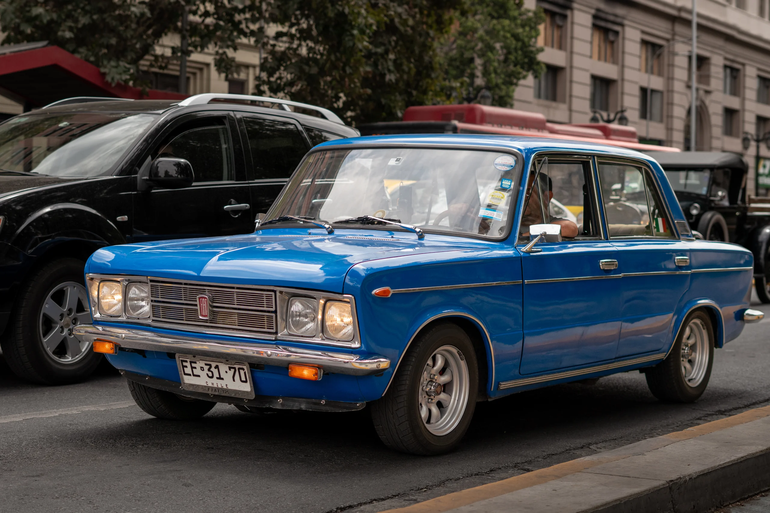 Imagen de un automóvil clásico Fiat en color azul estacionado o detenido en una calle urbana con edificios y otros vehículos alrededor. El auto tiene placa chilena y detalles vintage en su diseño, reflejando un estilo de vehículo de épocas pasadas.