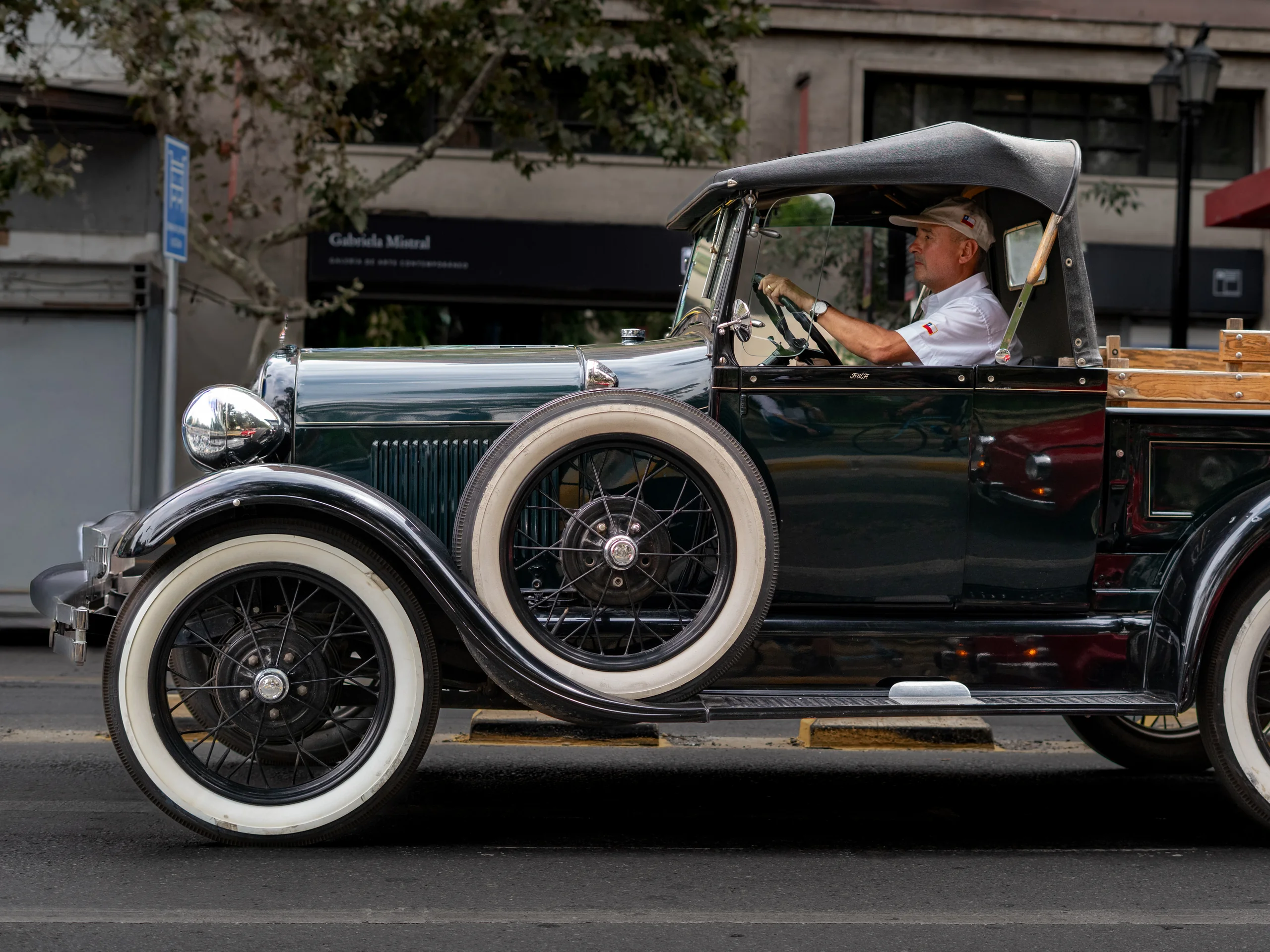 En la imagen se observa un vehículo antiguo tipo camioneta con llantas de borde blanco, conducido por un hombre que viste una camiseta blanca y gorra beige. El vehículo está detenido o circulando en una calle urbana con edificios y árboles en el fondo.