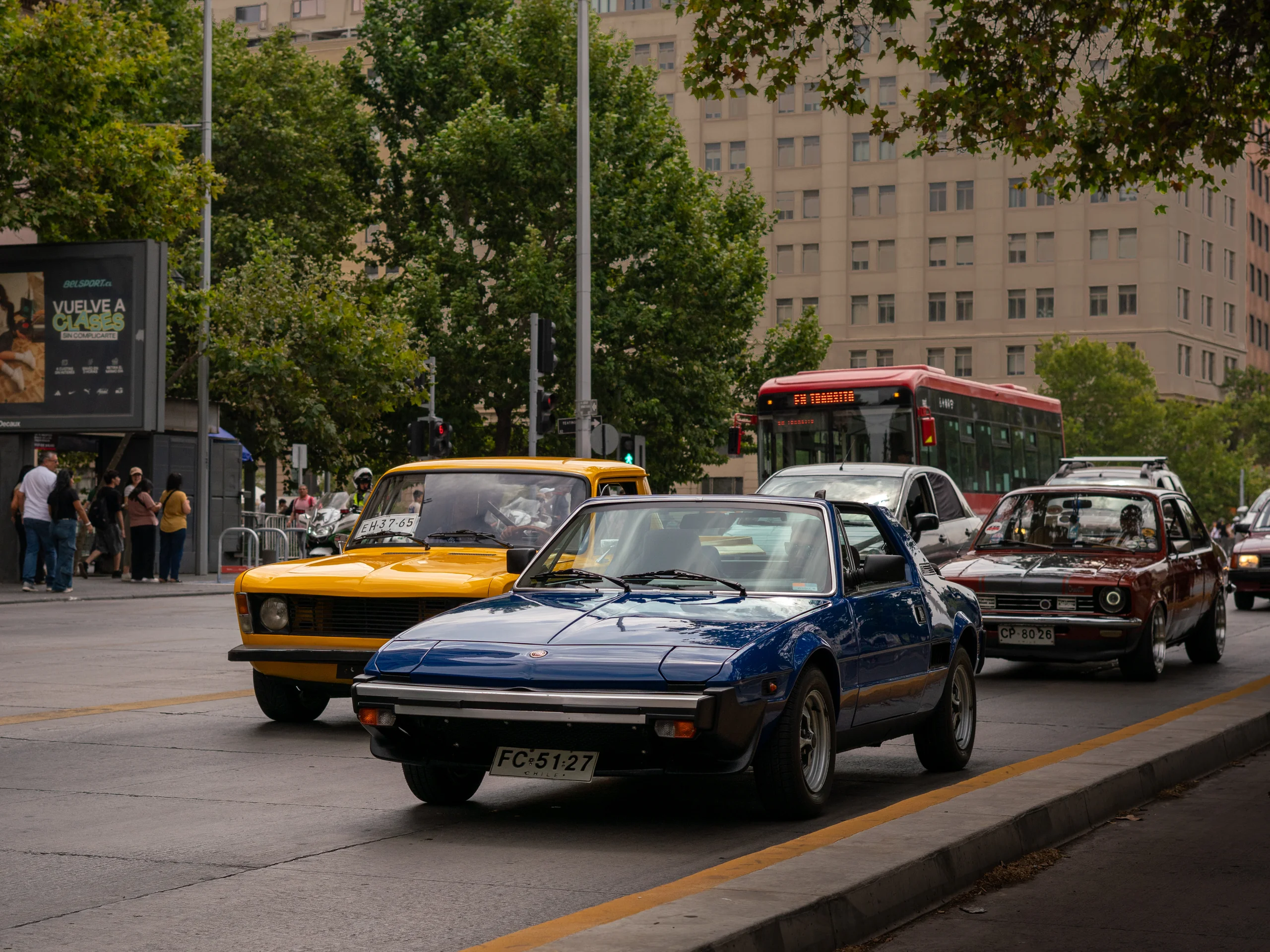 La imagen muestra una calle urbana con varios autos clásicos detenidos o circulando. Al frente hay un automóvil deportivo azul, seguido por un auto amarillo, otro carro rojo y un bus rojo y blanco al fondo. Del lado izquierdo se observan peatones esperando en la esquina de la calle, mientras que edificios altos y árboles rodean la escena, indicando un entorno metropolitano.