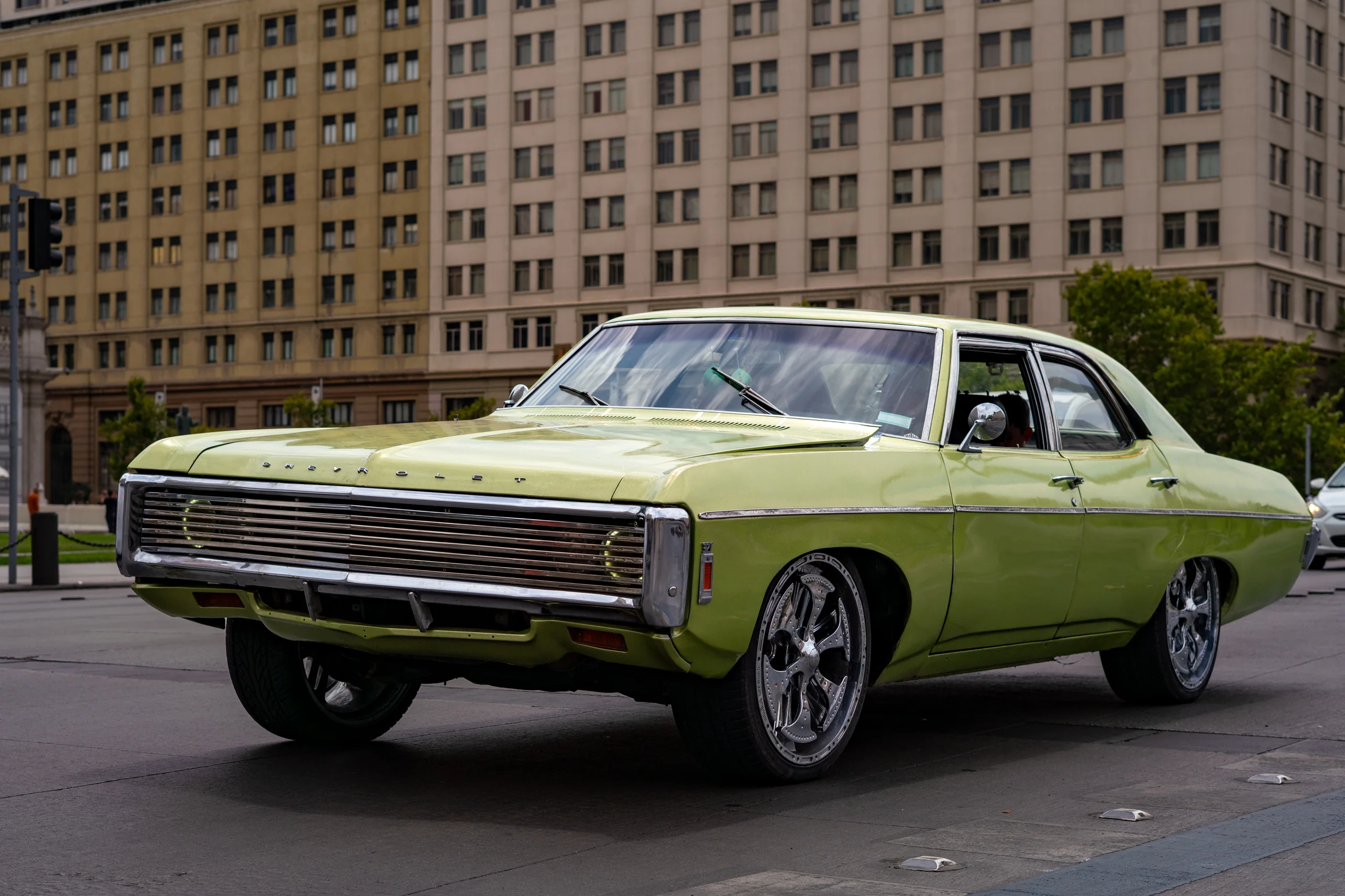 Automóvil clásico de color verde lima con detalles cromados, estacionado en una calle pavimentada en una zona urbana, con edificios altos de fondo. El vehículo tiene un diseño de los años 70 y se observa bien conservado, con llantas modernas y detalles brillantes en la carrocería.