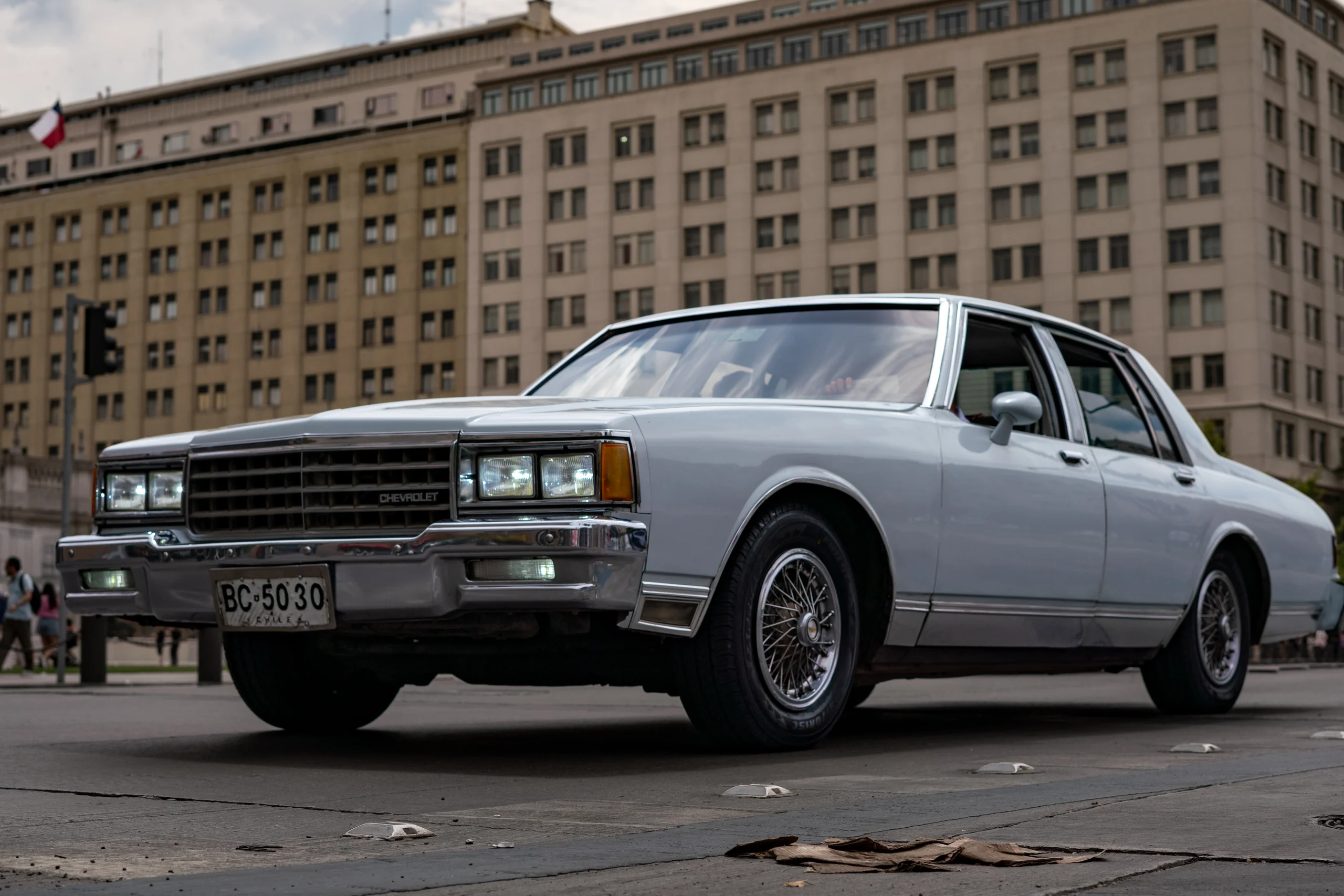 Vehículo clásico Chevrolet de color blanco estacionado o circulando sobre una calle asfaltada en un entorno urbano, con un edificio alto y antiguo de fondo. La escena muestra un día nublado o con luz difusa, y detalles del auto como las llantas y luces delanteras están claramente visibles.