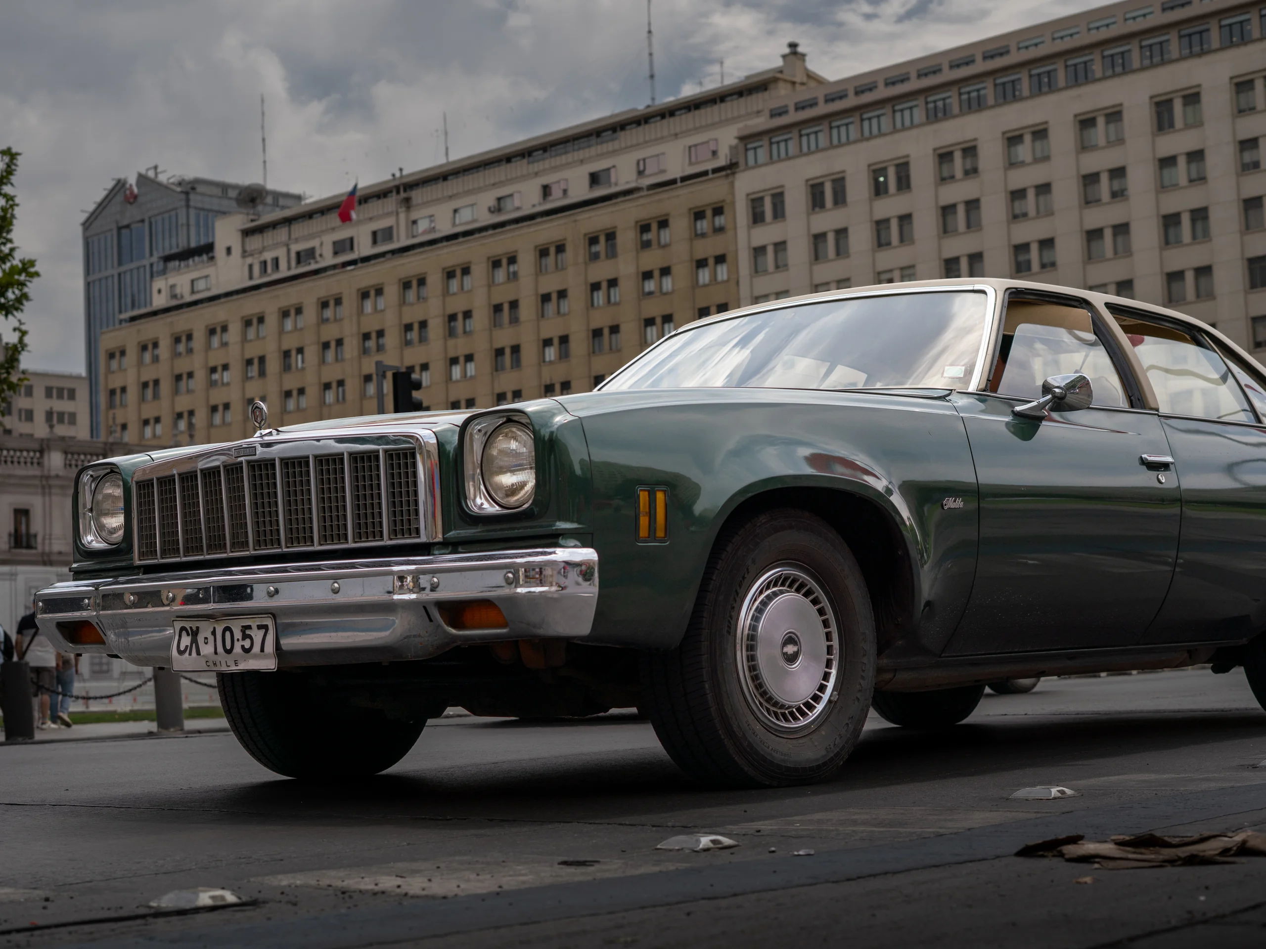 Un automóvil clásico de color verde oscuro estacionado sobre una calle asfaltada, con un edificio grande de varios pisos y ventanas al fondo. El auto tiene guardabarros cromados y matrícula visible con la inscripción 'Chile'. El cielo está nublado, lo que proporciona una luz suave a la escena.