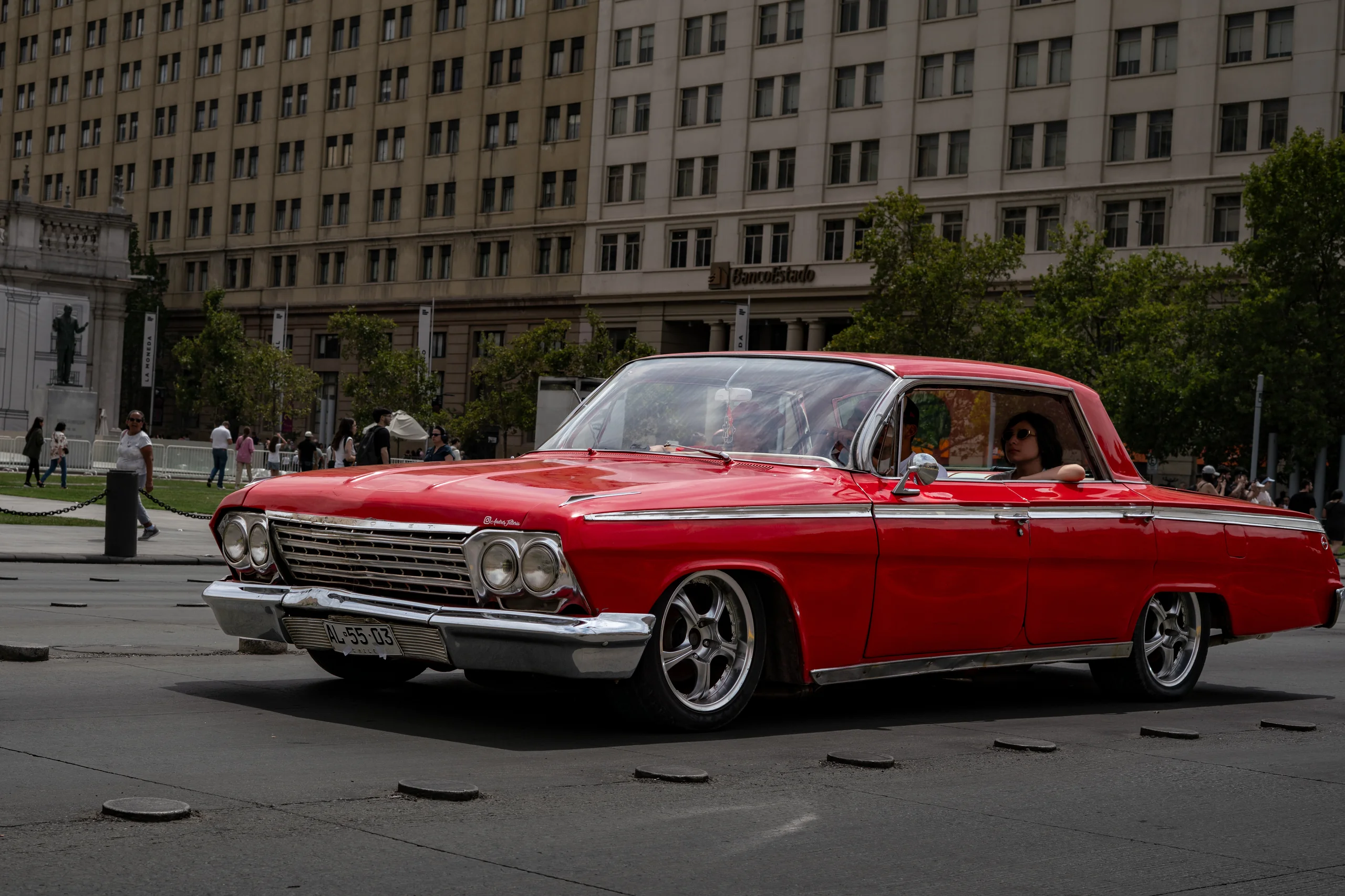Imagen de un automóvil clásico rojo circulando por una calle de una ciudad con edificios de arquitectura tradicional de fondo. El auto lleva una persona con gafas de sol en el asiento del conductor. Alrededor se observan más personas caminando y una zona arbolada junto a la acera.