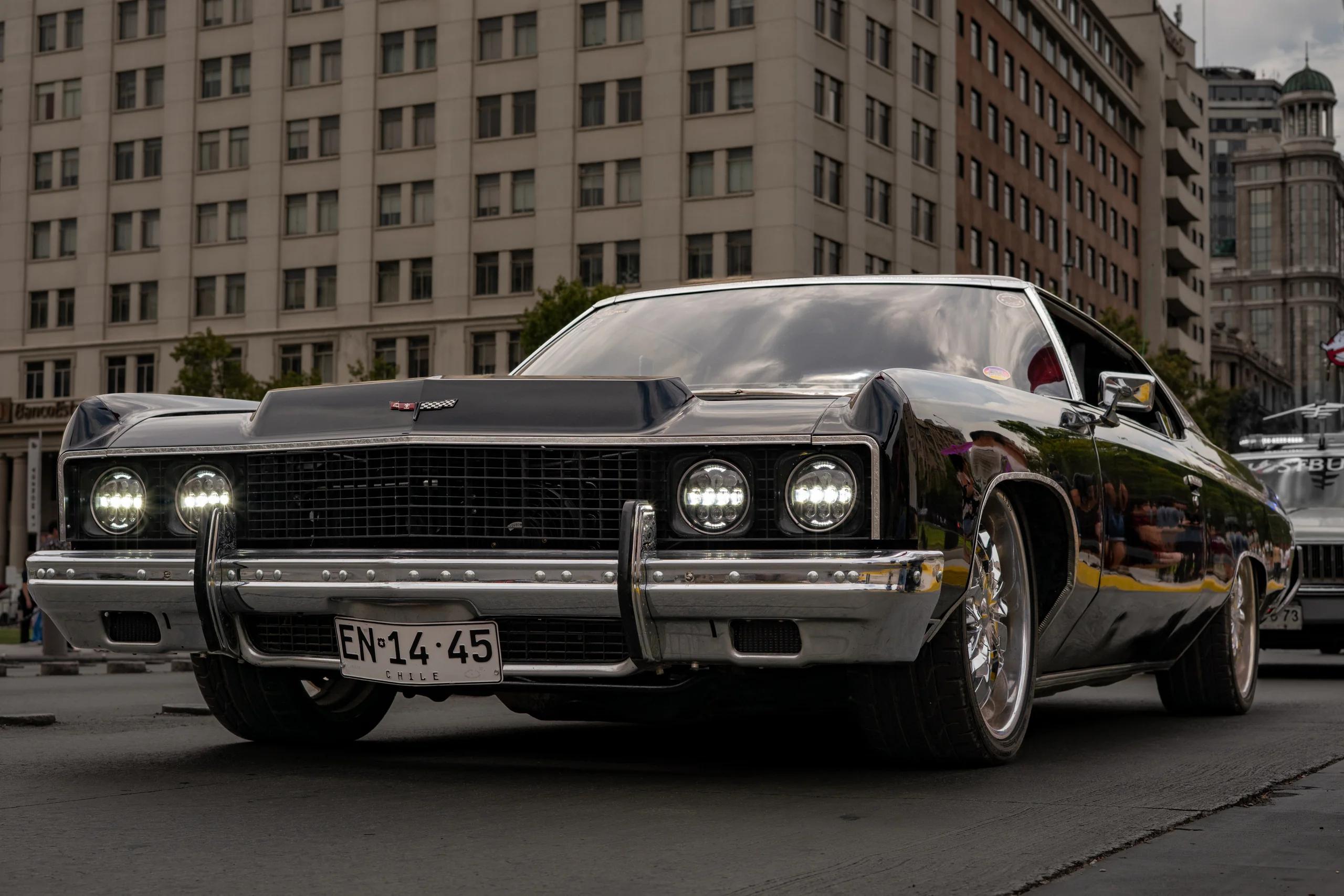 Automóvil clásico negro con detalles cromados y luces delanteras encendidas, estacionado en una calle urbana con edificios altos y arquitectura moderna al fondo. El vehículo tiene placa patente chilena y su carrocería refleja el entorno cercano.