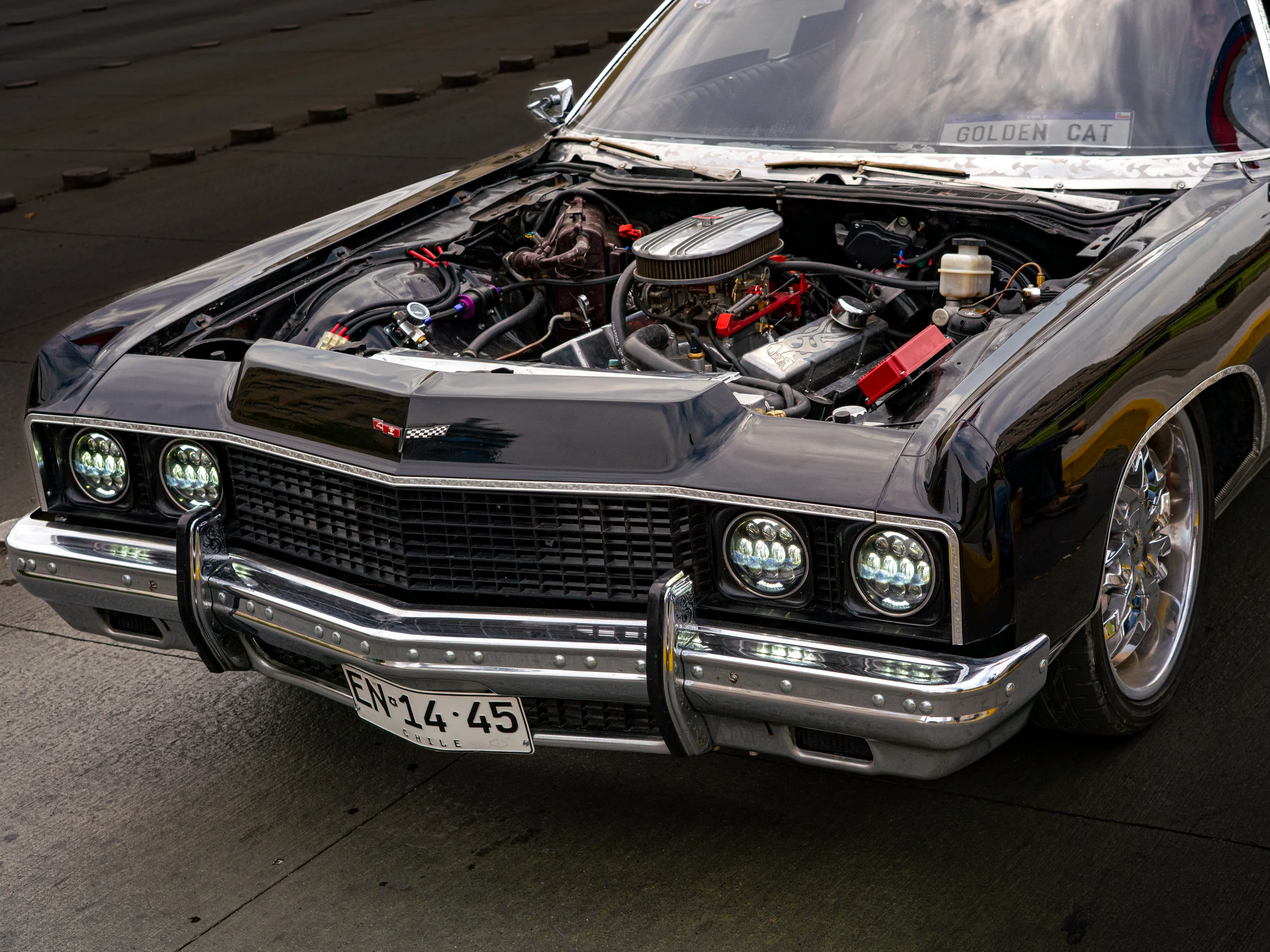 Se muestra un automóvil clásico de color negro estacionado con el capó abierto, revelando un motor cromado y detalles mecánicos visibles. El vehículo tiene faros delanteros modernos LED y una placa patente chilena visible en la parte frontal.