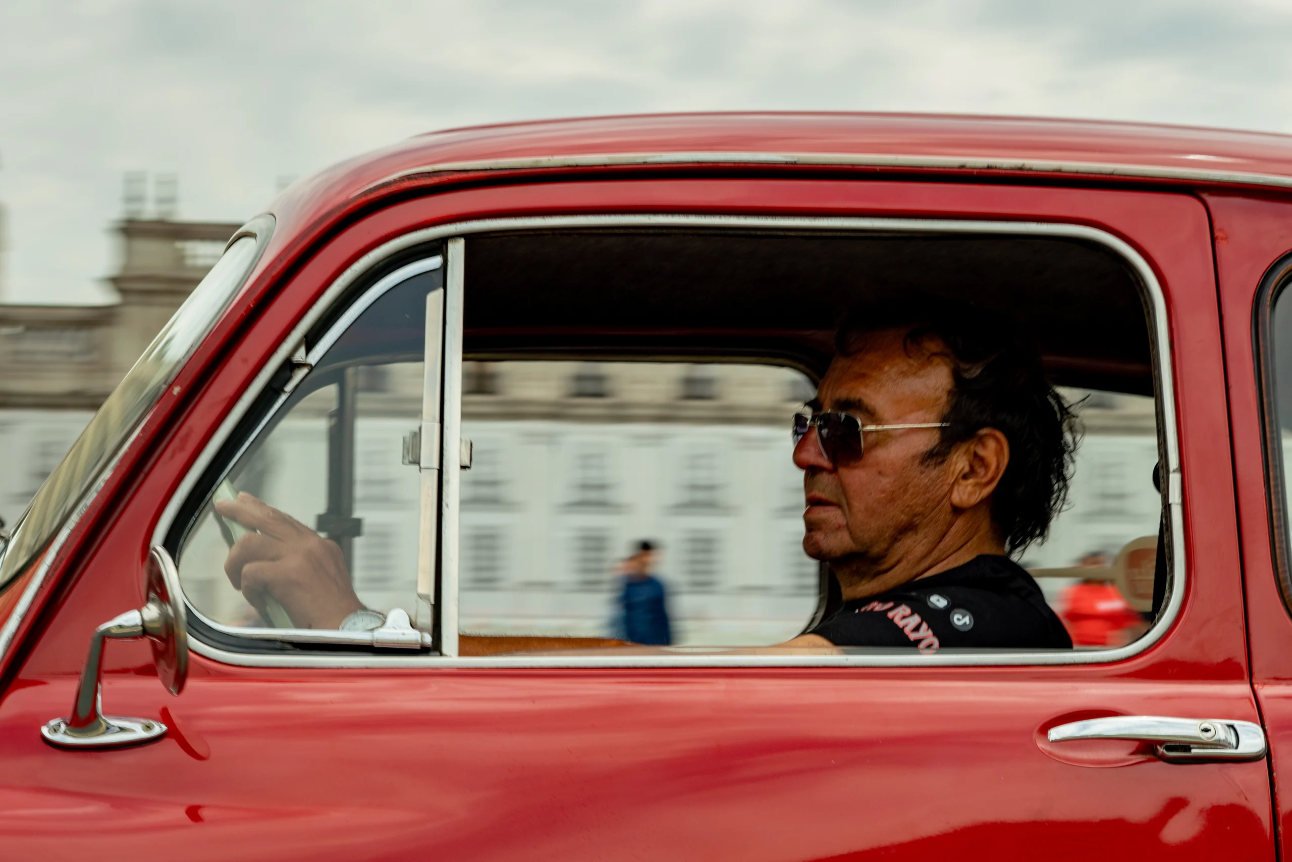 Un hombre mayor con gafas oscuras está conduciendo un vehículo clásico de color rojo. La imagen muestra un perfil lateral del conductor, con el fondo desenfocado que sugiere un entorno urbano.