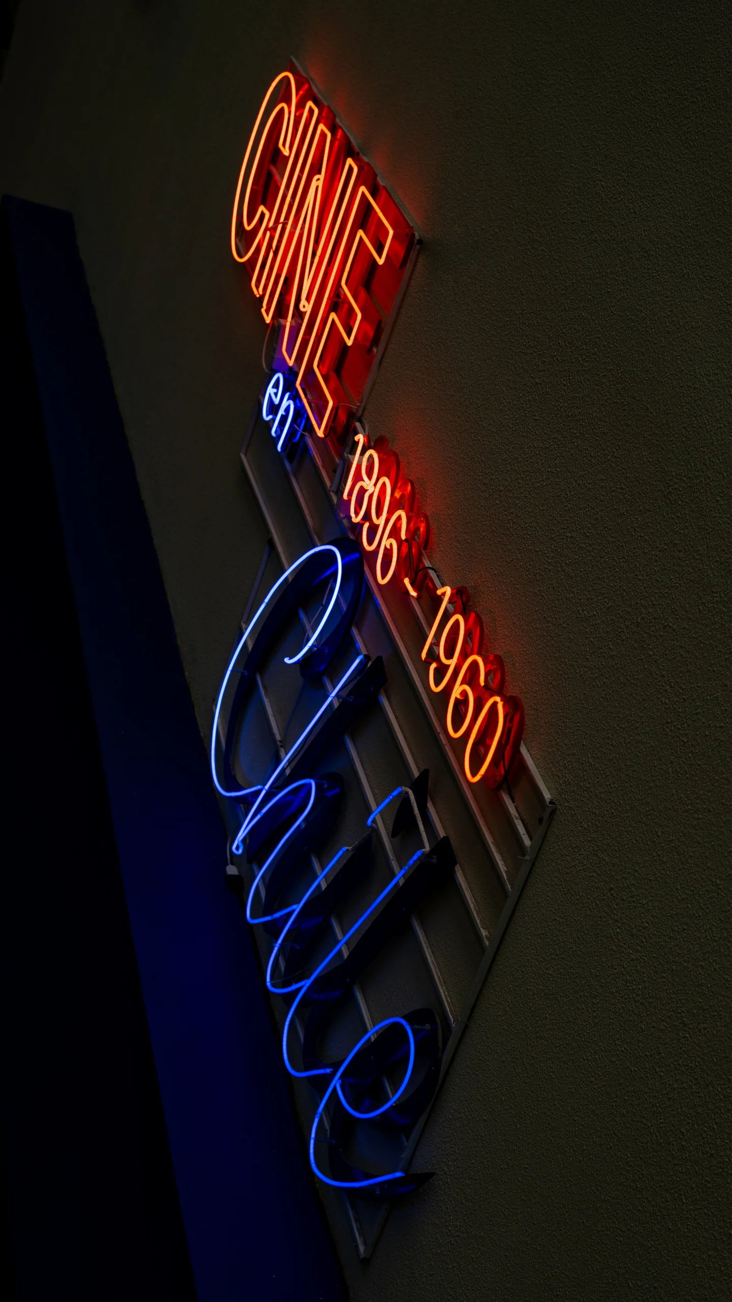 La imagen muestra un cartel de neón colocado verticalmente en una pared. El texto en el cartel está dividido en tres colores: la palabra CINE en rojo, la palabra en en azul y Chile en azul eléctrico. Además, las fechas 1896-1960 están en rojo, indicando un posible homenaje o exposición relacionada con la historia del cine en Chile dentro de ese rango de años.