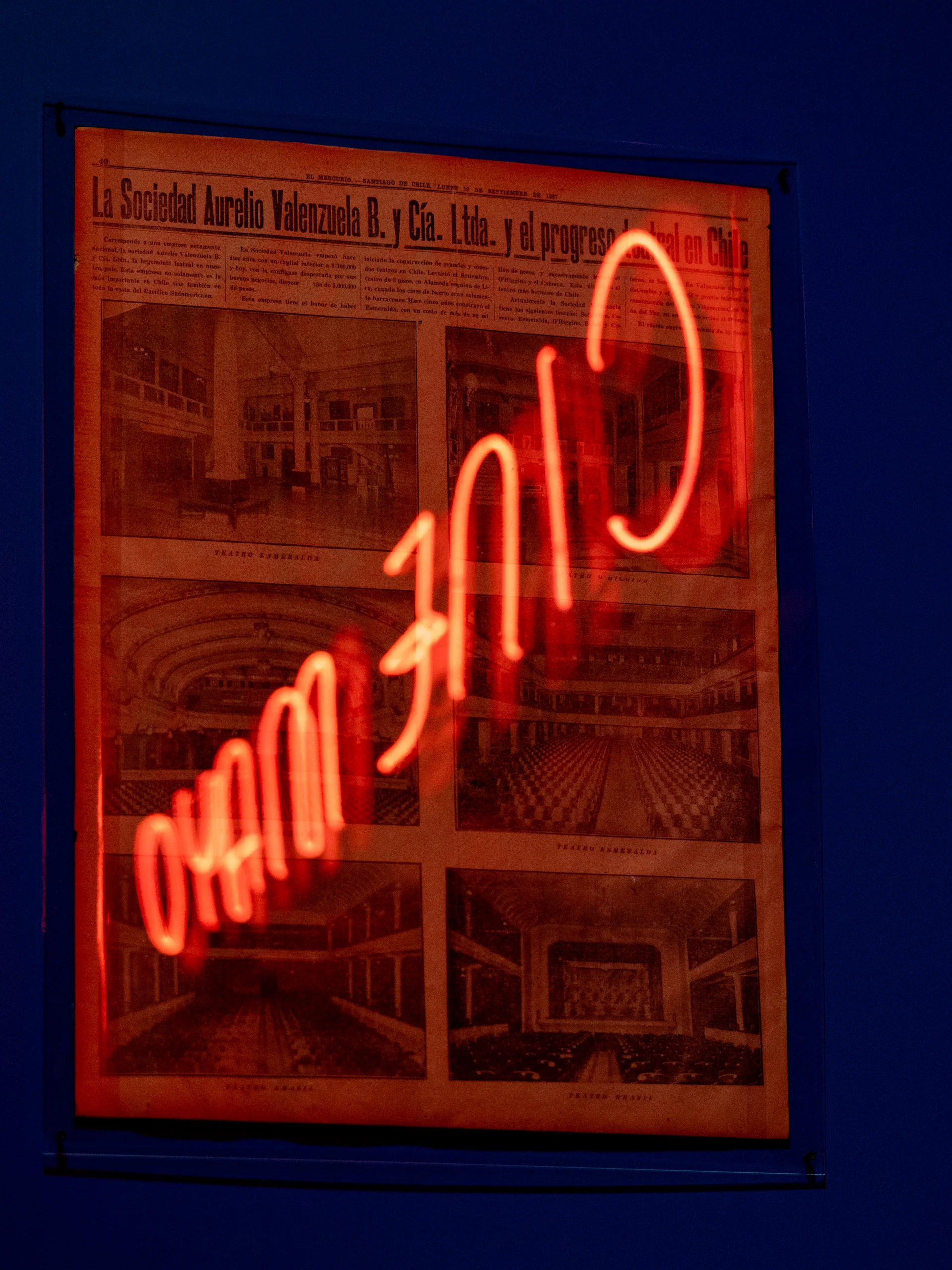 La imagen muestra un cartel antiguo con texto en español que habla de la Sociedad Aurelio Valenzuela B. y Cía. Ltda. y el progreso teatral en Chile. Incluye varias fotografías en blanco y negro de teatros interiores. Sobre el cartel hay un reflejo o proyección de un letrero en neón rojo que parece un conjunto de números.