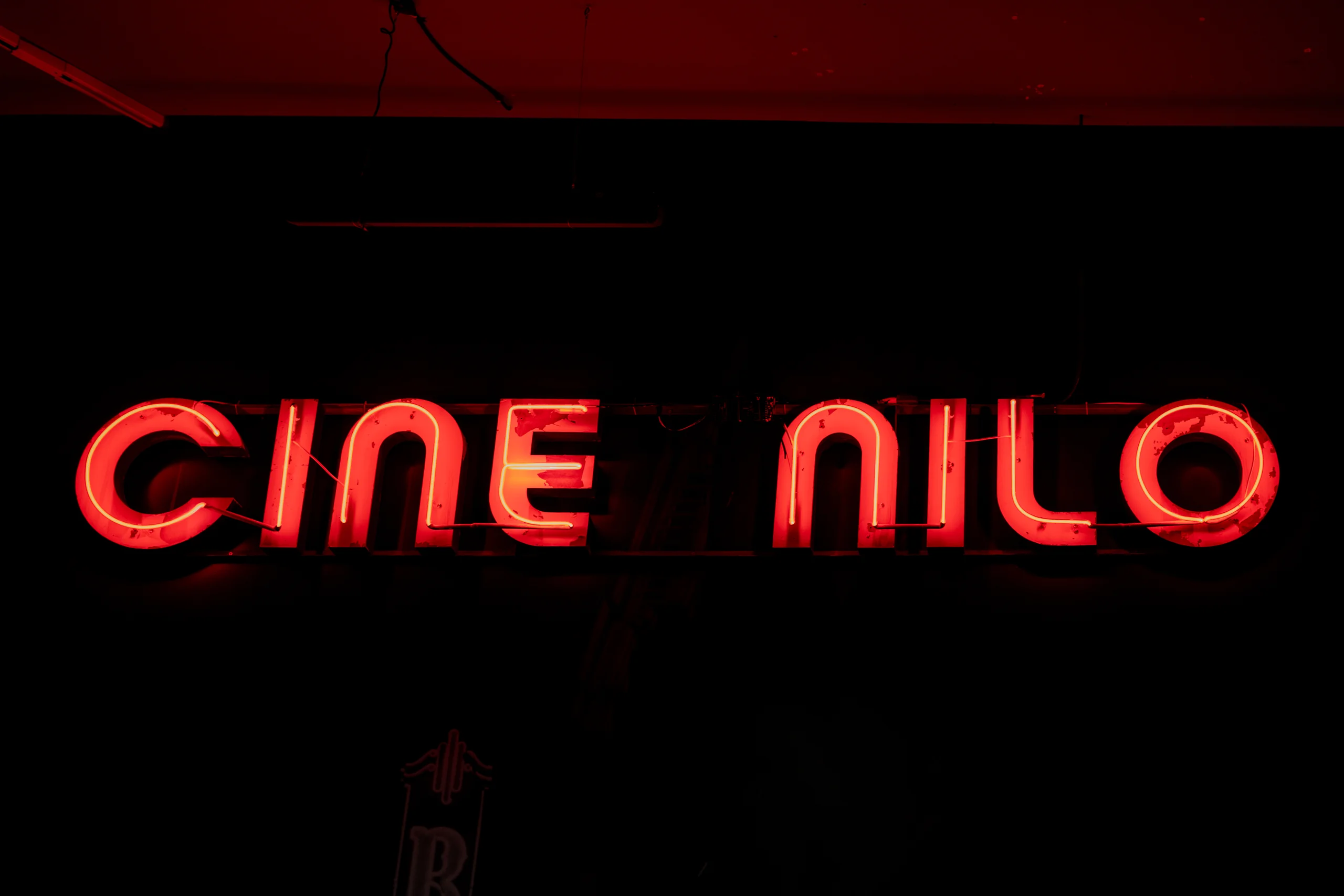 La imagen muestra un cartel luminoso de neón de color rojo con el texto 'Cine Nilo'. Está colocado sobre un fondo oscuro y las letras destacan claramente por el brillo del neón. El cartel parece pertenecer a un cine o sala de proyección llamada 'Nilo'.