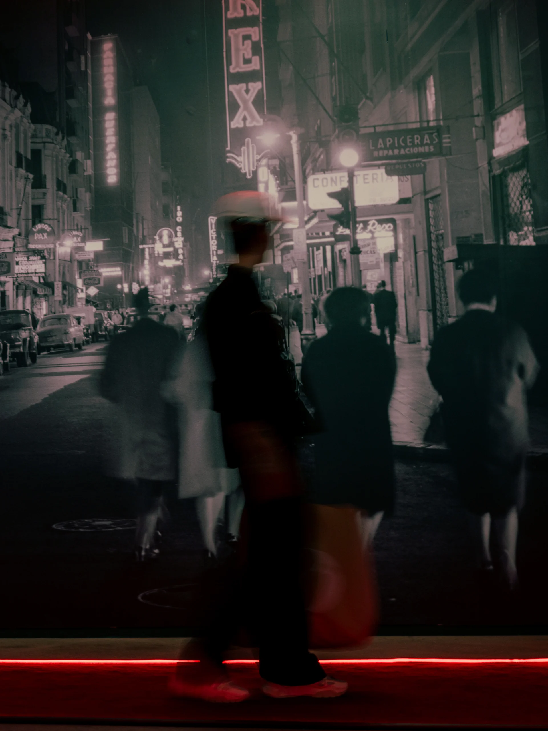 Imagen en blanco y negro de una calle nocturna en una ciudad con varios letreros luminosos visibles. Se observan personas caminando, algunas con vestimenta de época, con un efecto de movimiento que crea desenfoque. En primer plano, una figura borrosa en colores parcialmente visibles, caminando sobre una línea de luz roja en el suelo.