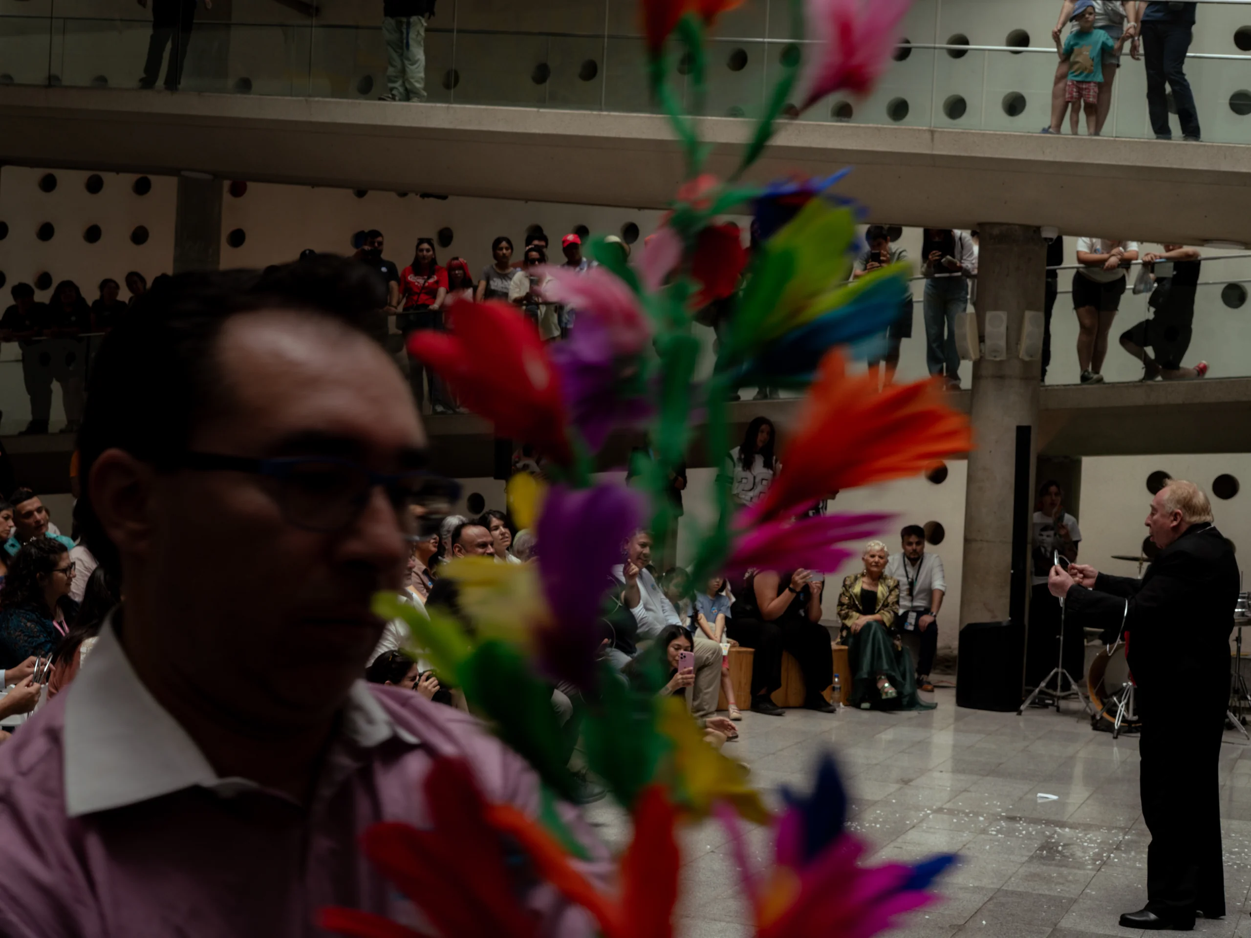 La imagen muestra a un hombre mayor vestido de negro tocando un instrumento, posiblemente una trompeta o similar, en un espacio cerrado con una audiencia sentada y de pie observando. Hay personas en dos niveles del lugar y en primer plano se observa desdibujada una planta o adorno con flores de colores brillantes que ocupa gran parte del encuadre frente a la cámara.