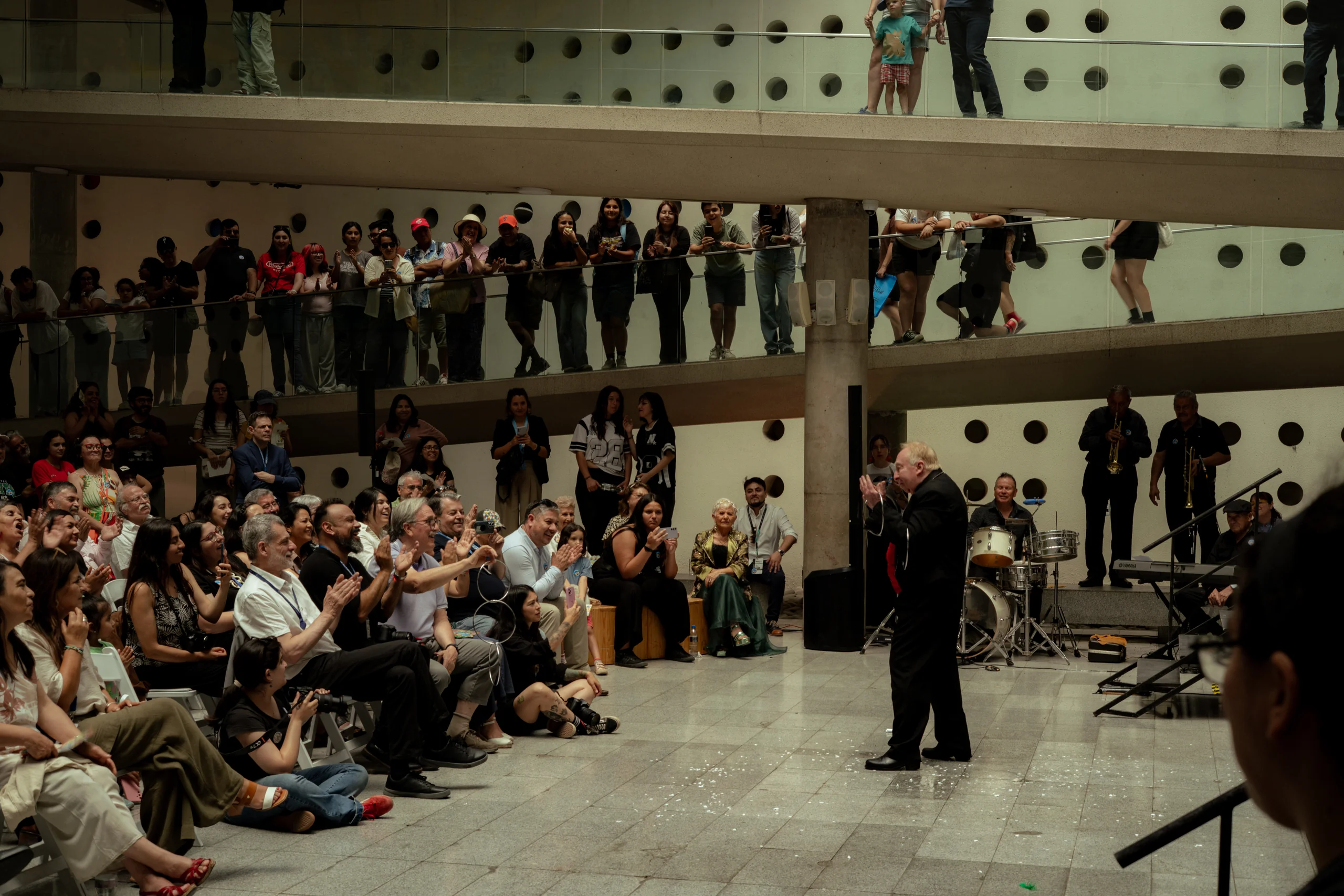 Un evento cultural o artístico en un espacio cerrado con dos niveles. En el nivel inferior, un hombre de edad avanzada vestido de negro se encuentra frente a un público que lo observa y aplaude, mientras que en el fondo hay músicos tocando instrumentos como trompeta, batería y teclado. En el segundo nivel, varias personas están de pie observando el evento desde un balcón con barandales de vidrio y una pared decorada con círculos.